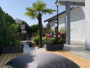 A patio with a wooden deck, a grey cushion, potted plants, and a palm tree. - 5-Room Villa with Pool and Terrace in Le Porge (Le Porge)