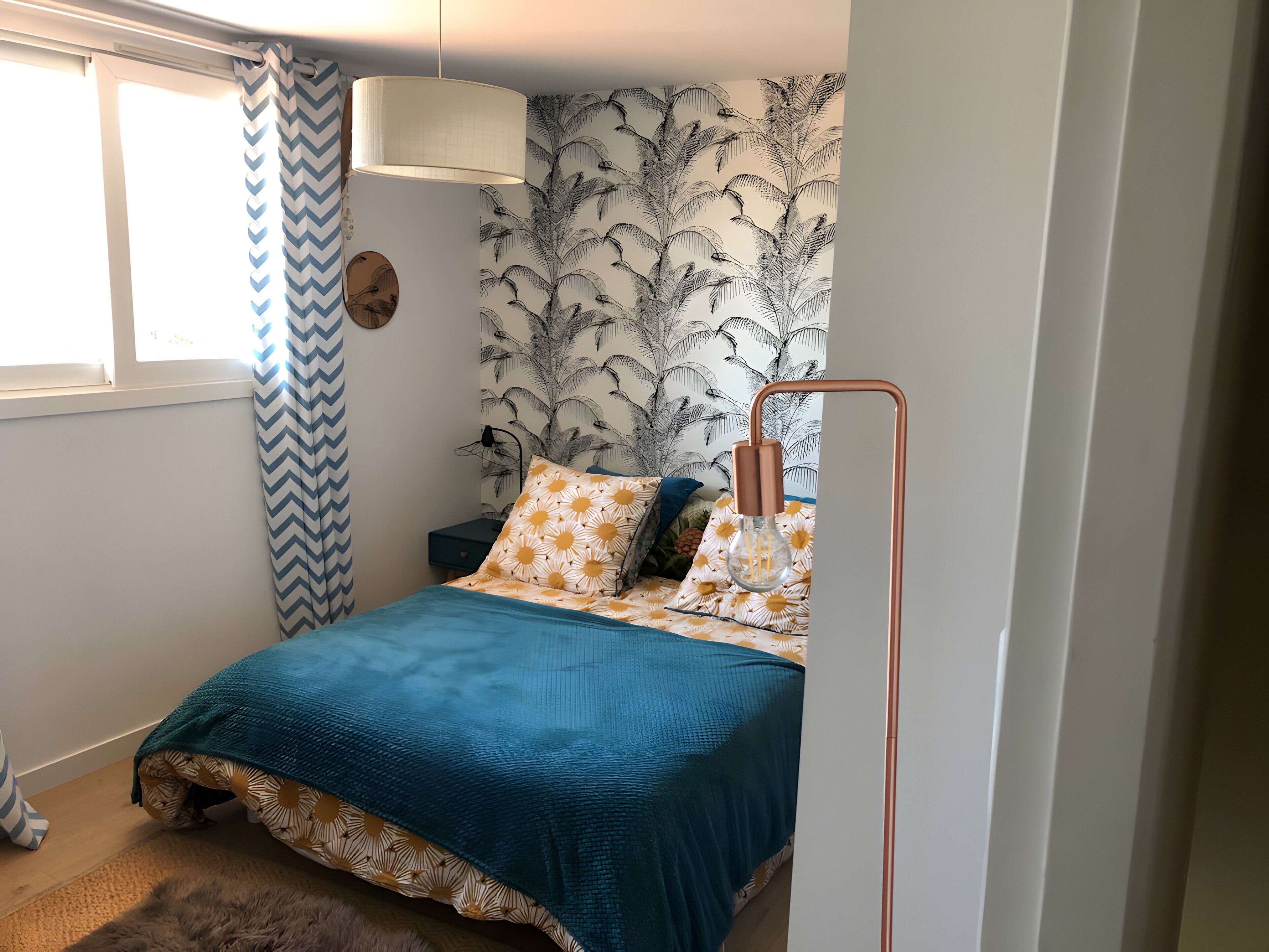 Ein Schlafzimmer mit Bett, Nachttisch, Lampe, Fenster mit Vorhängen und einer Wand mit Blattmuster.