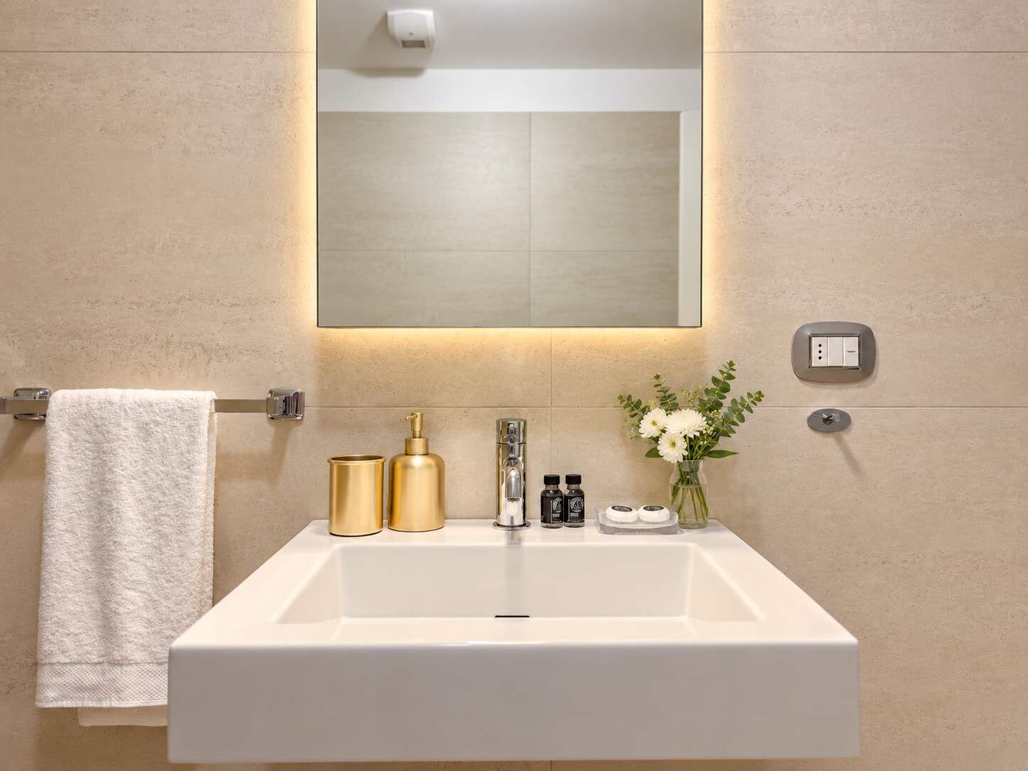 Un baño con un lavabo blanco, dispensadores de jabón dorados, un espejo y un perchero para toallas.