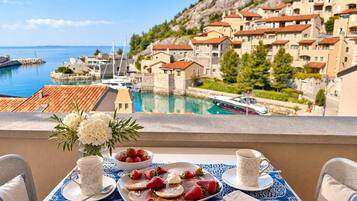 Un balcone con un tavolo apparecchiato per la colazione, con vista su una cittadina costiera e edifici storici e un porto turistico.