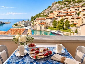 A balcony with a table set for breakfast, overlooking a coastal town with historic buildings and a marina. - Sole Mio - Portopiccolo Apartments (Duino Aurisina)