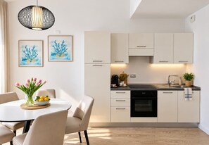 A modern kitchen with white cabinets, a black dishwasher, and a dining area with a table and chairs. - Sole Mio - Portopiccolo Apartments (Duino Aurisina)