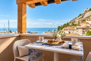A balcony with a table set for two, overlooking a coastal view with buildings and the sea. - Sole Mio - Portopiccolo Apartments (Duino Aurisina)