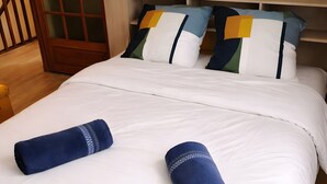 Un letto rifatto con biancheria bianca e due asciugamani blu arrotolati, un quadro raffigurante un felce sulla testiera e una porta in legno con un pannello di vetro.