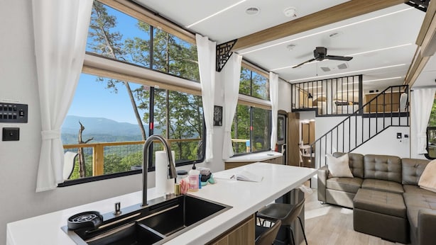 A modern kitchen with a large window, a dining area, and a living room with a sofa. - The Chestnut- Smoky Mountains Tiny House w/ Hot Tub & Lake (Tallassee)