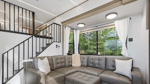 A modern living room with a sectional sofa, a staircase, and large windows. - The Chestnut- Smoky Mountains Tiny House w/ Hot Tub & Lake (Tallassee)