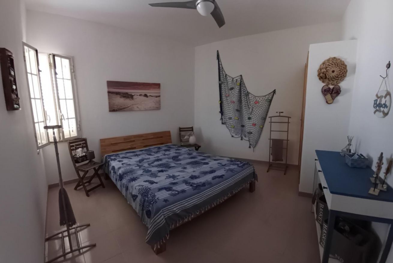 A bedroom with a bed, a standing fan, a chair, a desk, and a window.