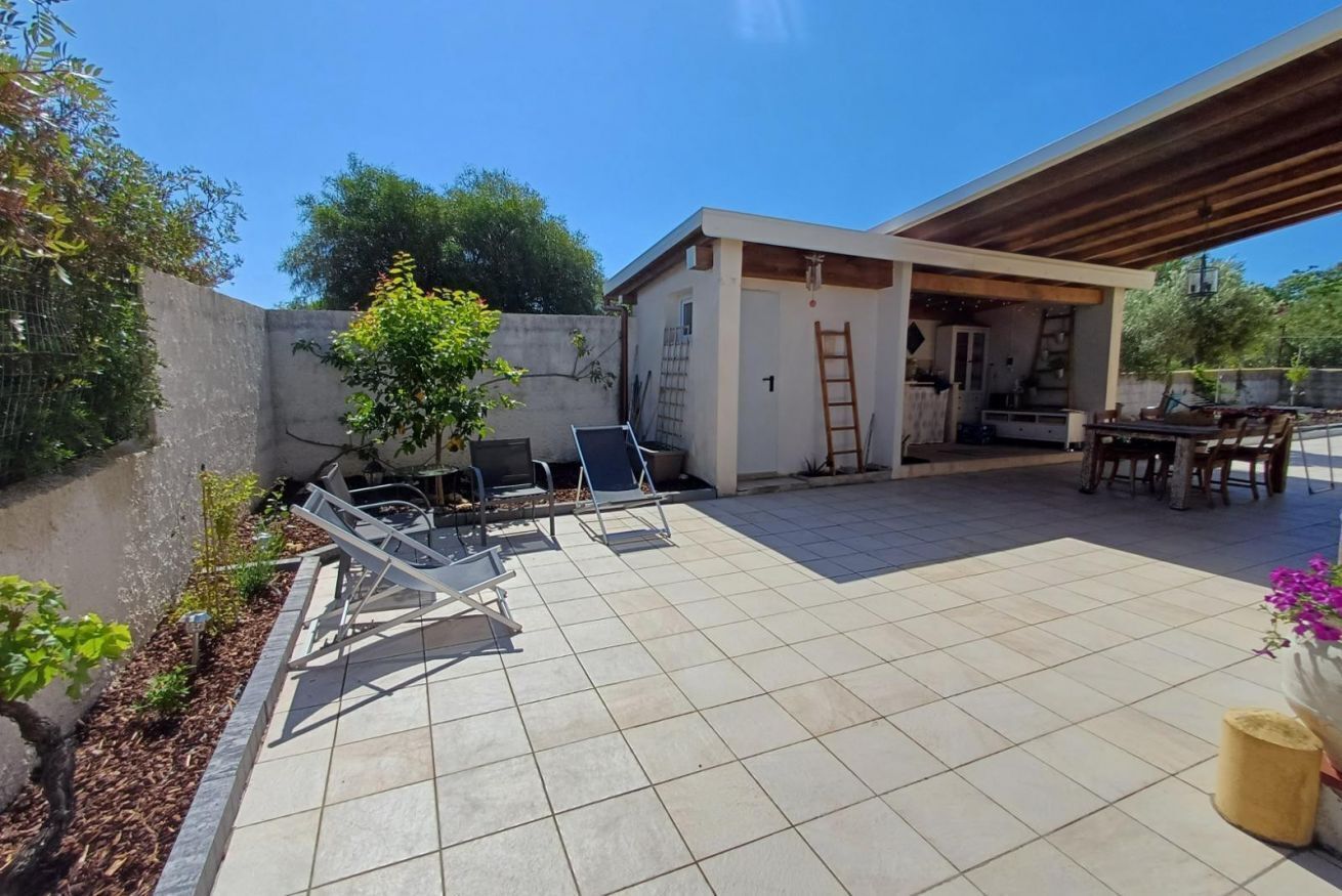 Een terras met betegelde vloer, buitenmeubilair, een klein gebouw en een overdekt gedeelte met een tafel en stoelen.