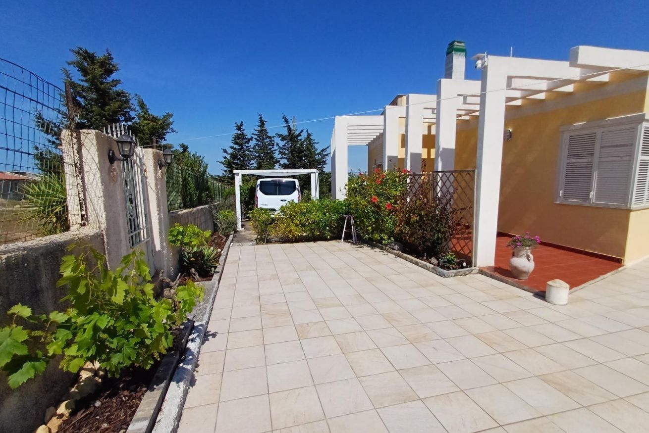 A paved patio with a white car parked in the background, surrounded by greenery and a white building.