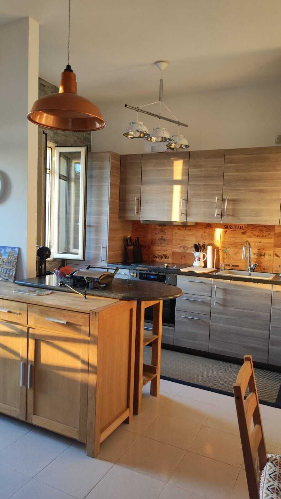 Ett modernt kök med träskåp, en köksö, hängande lampor och ett stort fönster.