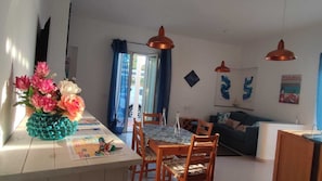 Ein Essbereich mit Tisch, Stühlen und einem Blumenstrauß. Im Hintergrund befinden sich eine Couch und eine Lampe.