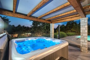Ein Whirlpool unter einer Holzw pergola mit Steinpfeilern, umgeben von einem angelegten Garten und einem Schwimmbad.