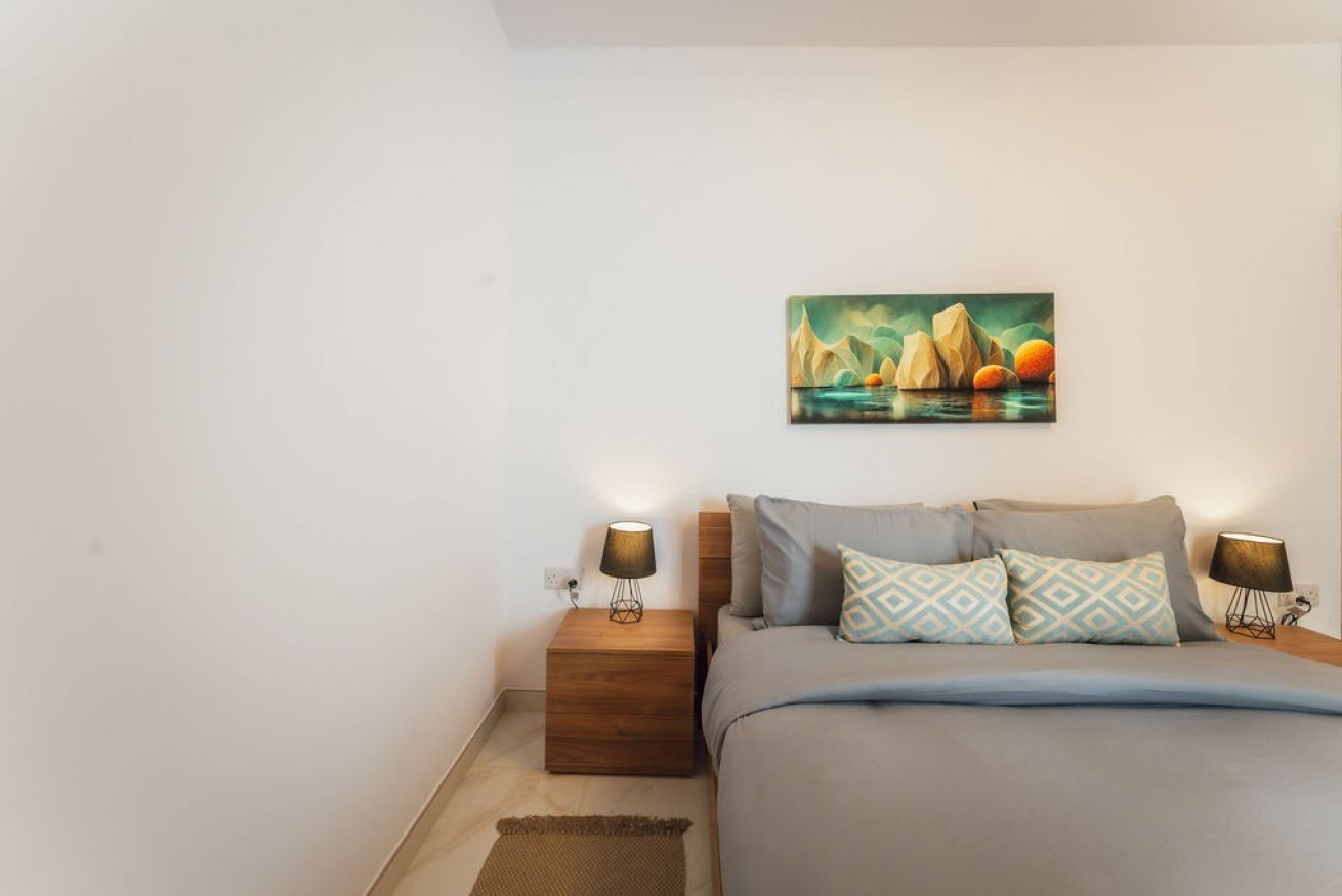 Quarto com cama, duas mesas de cabeceira, um criado-mudo, um quadro e uma luminária.