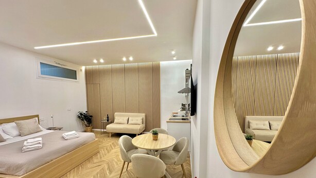 Quarto de hotel moderno com cama, sofá, mesa de jantar, cadeiras, espelho e área de cozinha.