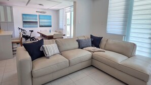 Una sala de estar moderna con un sofá seccional color beige, un comedor con mesa y sillas, y una cocina con armarios y encimera.