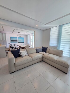 A modern living room with a beige sectional sofa, a dining area with a table and chairs, and a kitchen area with cabinets and a countertop. - New Remodeled 1BR· 2 Queen Beds+Full Kitchen+ Pool (Isabela)