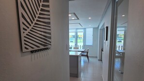 Un pasillo moderno con un cuadro de pared con motivos de hojas, un comedor con mesa y sillas, y vistas al exterior a través de amplias ventanas.