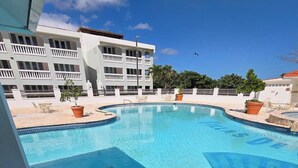Una piscina con plataforma de baldosas, un edificio blanco con balcones y un cielo despejado.