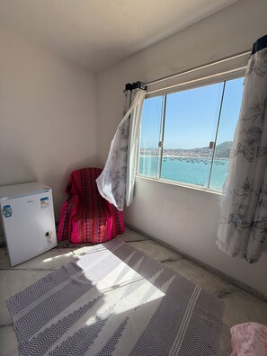 A room with a window overlooking a coastal view, a small white refrigerator, a patterned rug, and a red chair.