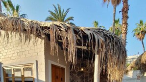A small, rustic building with a thatched roof, a stone wall, and a wooden door. There are wicker chairs and a small table outside.