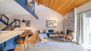 Een moderne woonkamer met een stenen open haard, een blauwe bank, een eethoek met een lange tafel en een trap met blauwe leuningen.