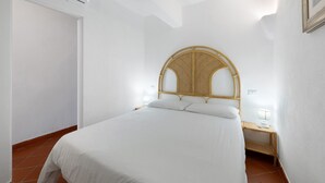 A single-bed room with a rattan headboard, white bedding, a bedside table with a lamp, and a wall-mounted air conditioning unit.