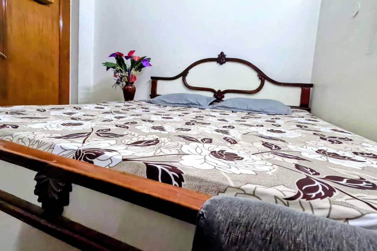 A bed with a floral-patterned bedspread, a wooden headboard, a nightstand with a vase of flowers, and a wooden door.