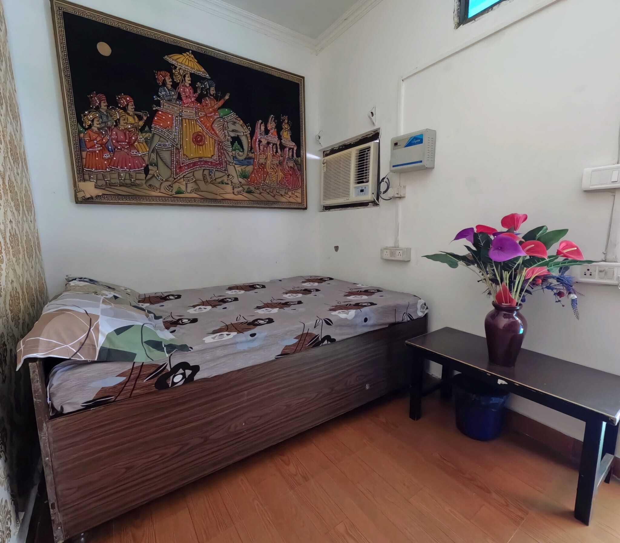 A room with a bed, a table, a vase with flowers, a wall-mounted air conditioner, and a painting.