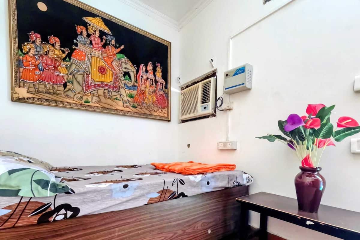 A room with a bed, a wall-mounted air conditioner, a painting, and a vase with flowers.