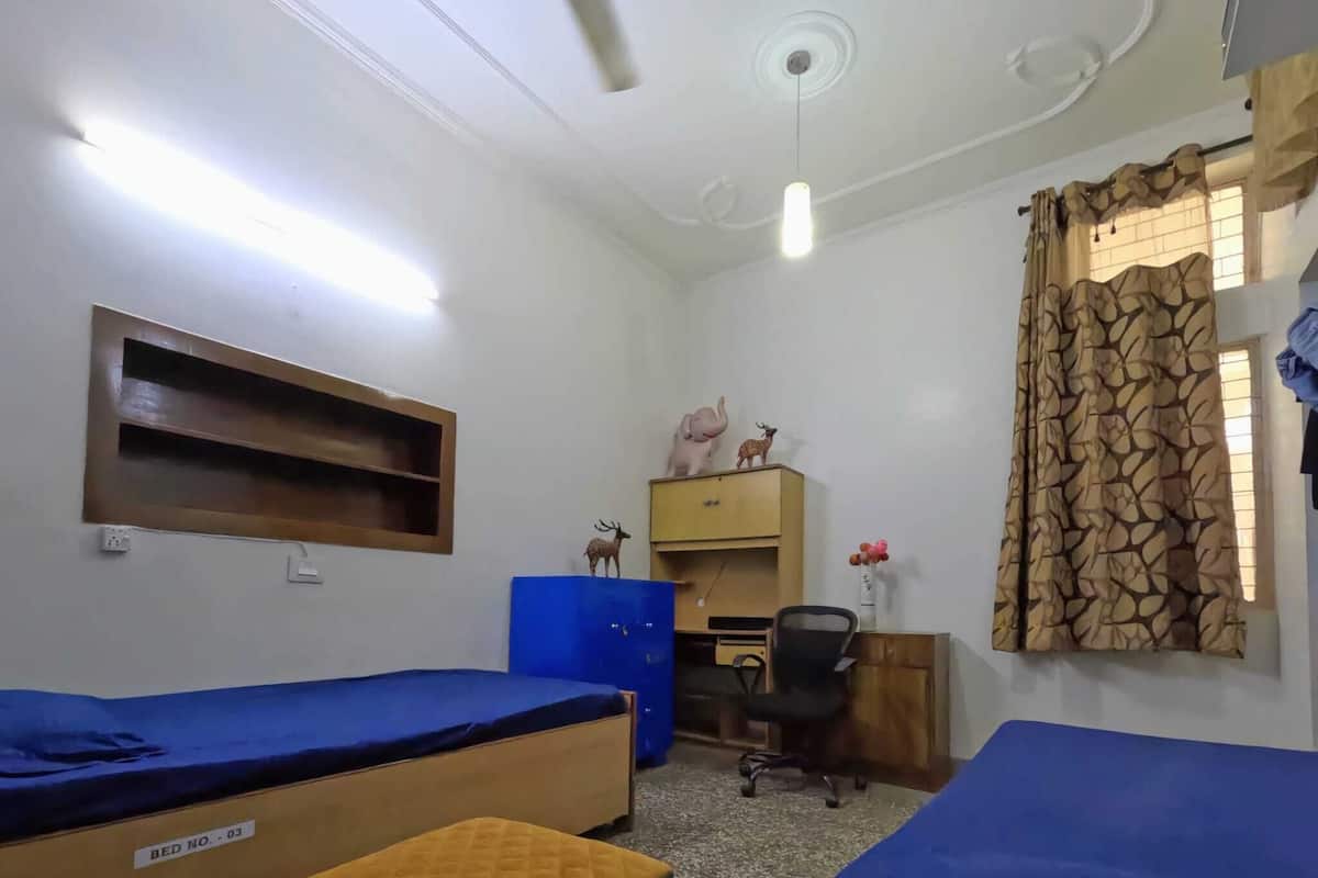 A dormitory room with two beds, a desk, a chair, a shelf with figurines, and a window with patterned curtains.