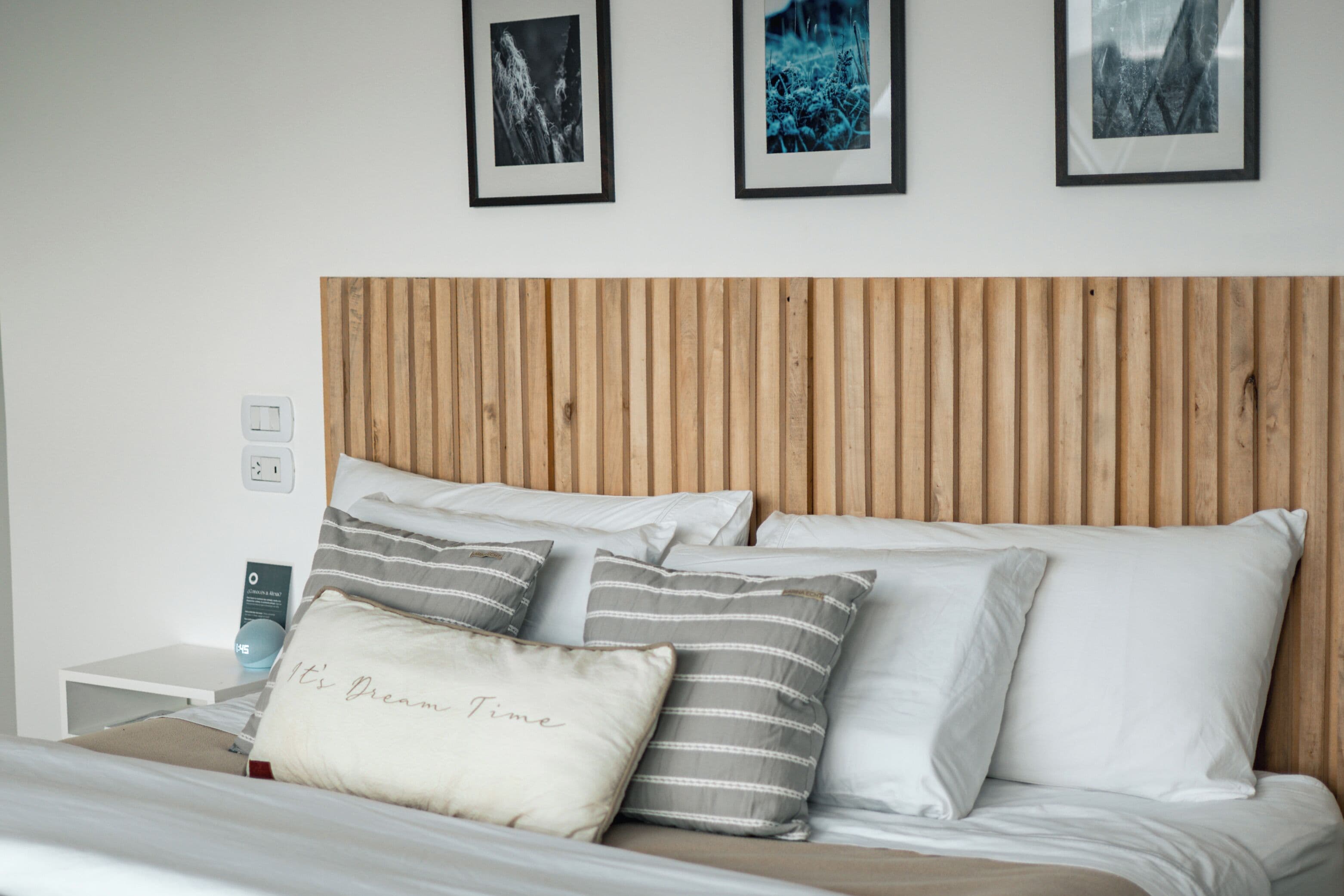 Una cama bien hecha con almohadas a rayas y lisas, cabecera de madera y cuadros enmarcados en la pared.