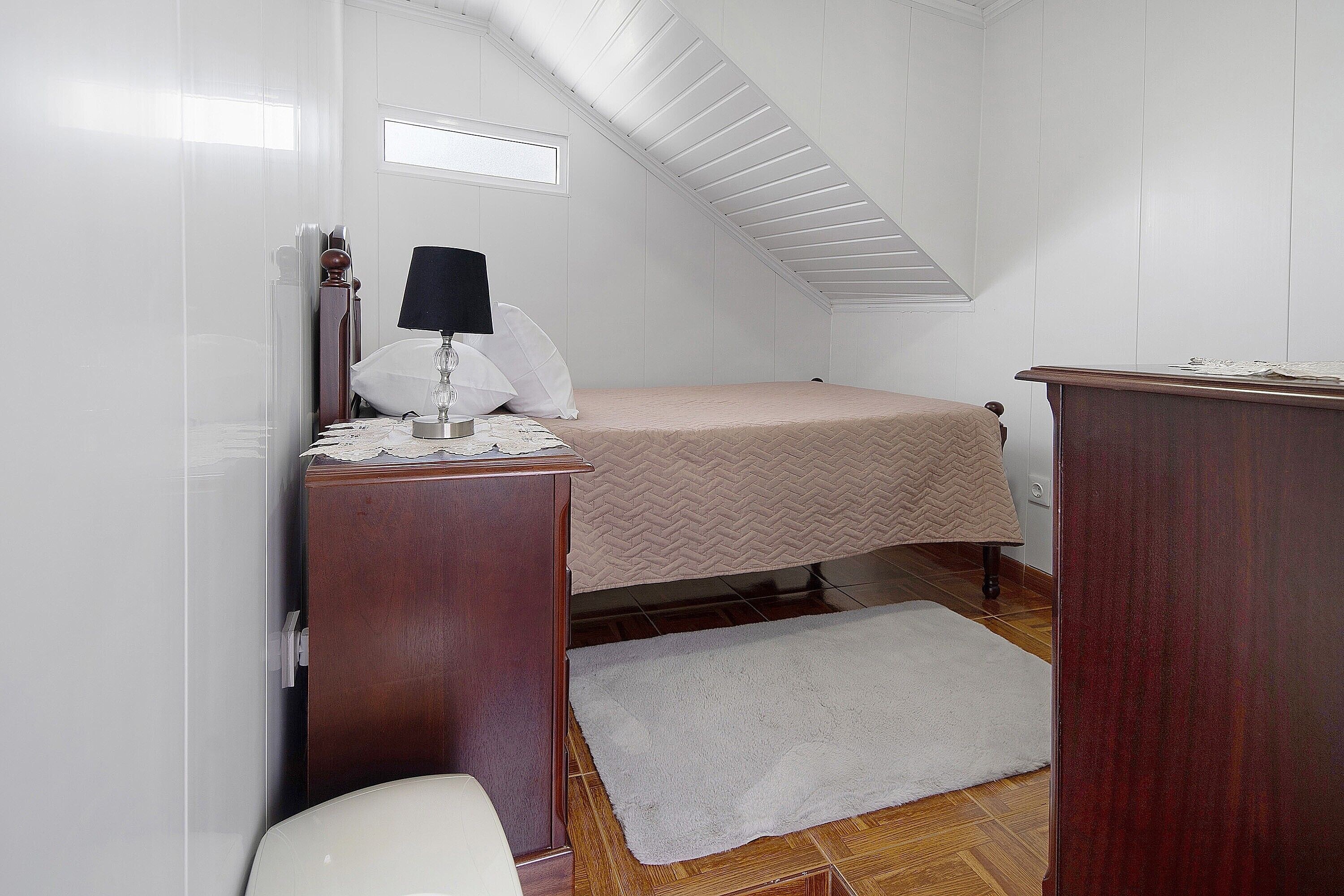 Une petite pièce bien éclairée, avec un lit, une commode en bois, un bureau avec une lampe et des toilettes blanches.