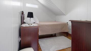 Une petite pièce bien éclairée, avec un lit, une commode en bois, un bureau avec une lampe et des toilettes blanches.
