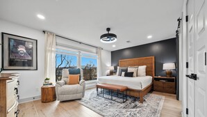 Ein Schlafzimmer mit einem großen Bett, einer Bank, einem Sessel, einem Nachttisch, einer Lampe, einer Kommode, einem gerahmten Bild, einem Fenster mit Vorhängen und einer Deckenleuchte.