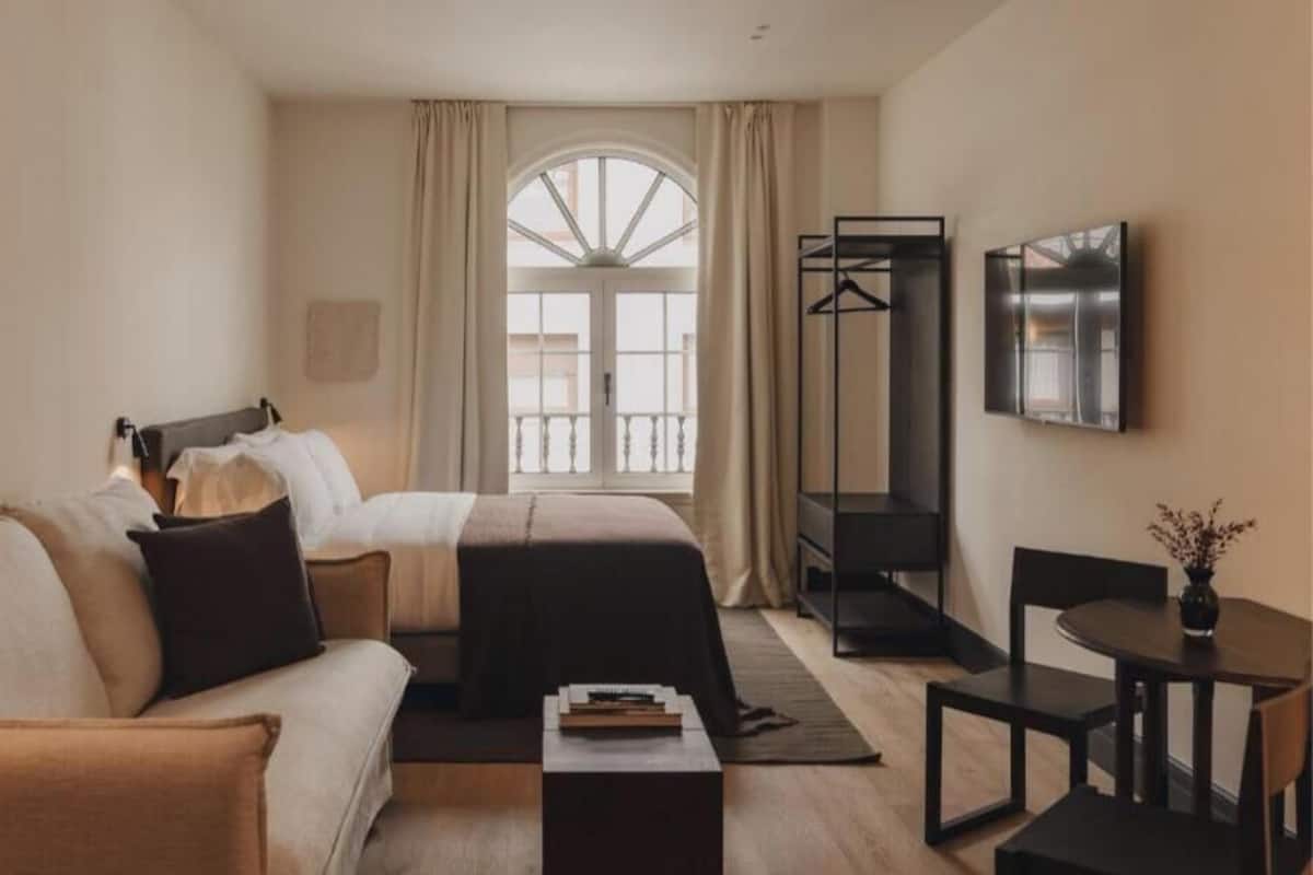 A hotel room with a bed, a sofa, a desk, a chair, a TV, and a window with curtains.