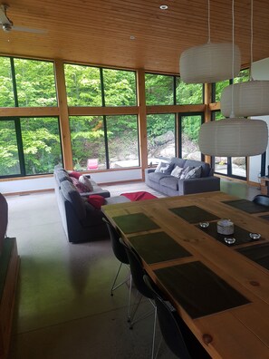 Ein modernes Wohnzimmer mit einem Holzesstisch, schwarzen Stühlen und einem grauen Sofa mit dekorativen Kissen. Große Fenster bieten Blick auf einen üppigen, grünen Wald.