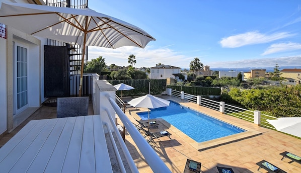 Une piscine entourée de chaises longues et de parasols, délimitée par une clôture blanche et donnant sur un quartier résidentiel avec des maisons et de la verdure.