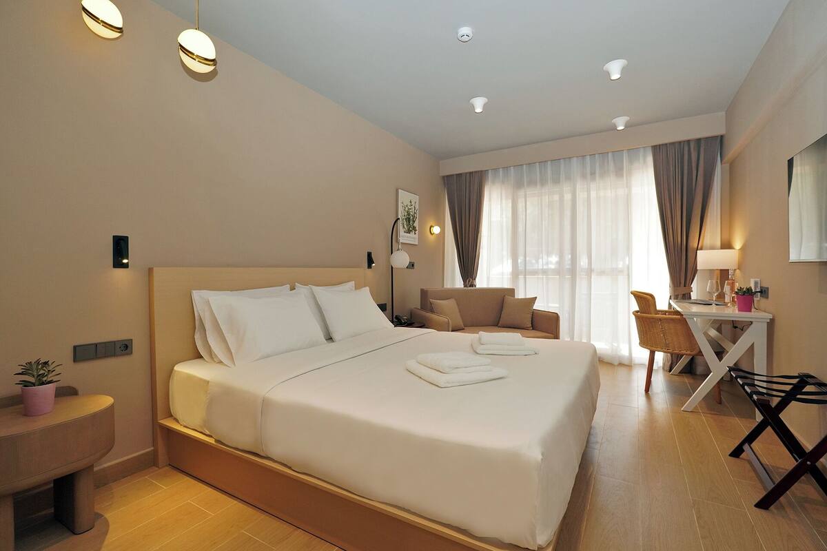 A hotel room with a large bed, a desk, a chair, and a small table with a lamp.