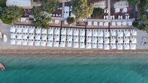 An aerial view of a beachside hotel with rows of sunbeds and umbrellas, a pool area, and a clear view of the turquoise sea.