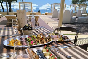 Une table à manger avec des assiettes remplies de nourriture, des verres de vin et des sièges d’extérieur protégés par des parasols.