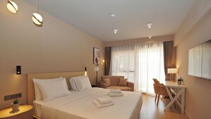 A hotel room with a large bed, a desk, a chair, a small table, a potted plant, and a window with curtains. - Ionian Crown Suites in Barbati  (Corfu)