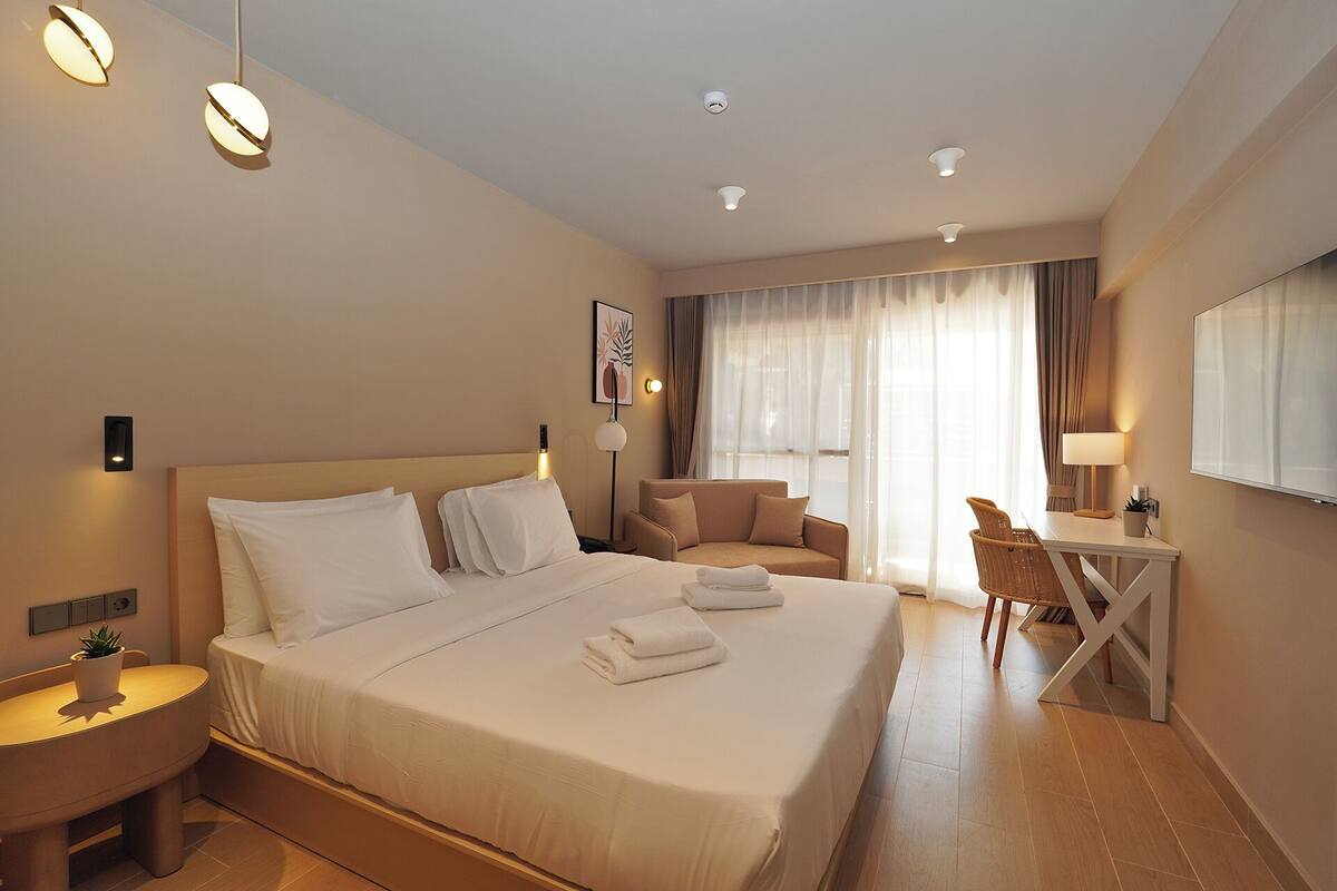A hotel room with a large bed, a desk, a chair, a small table, a potted plant, and a window with curtains.