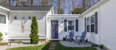 A white mobile home with a 'WELCOME' sign, a small porch with two chairs, and a pathway leading to the door.