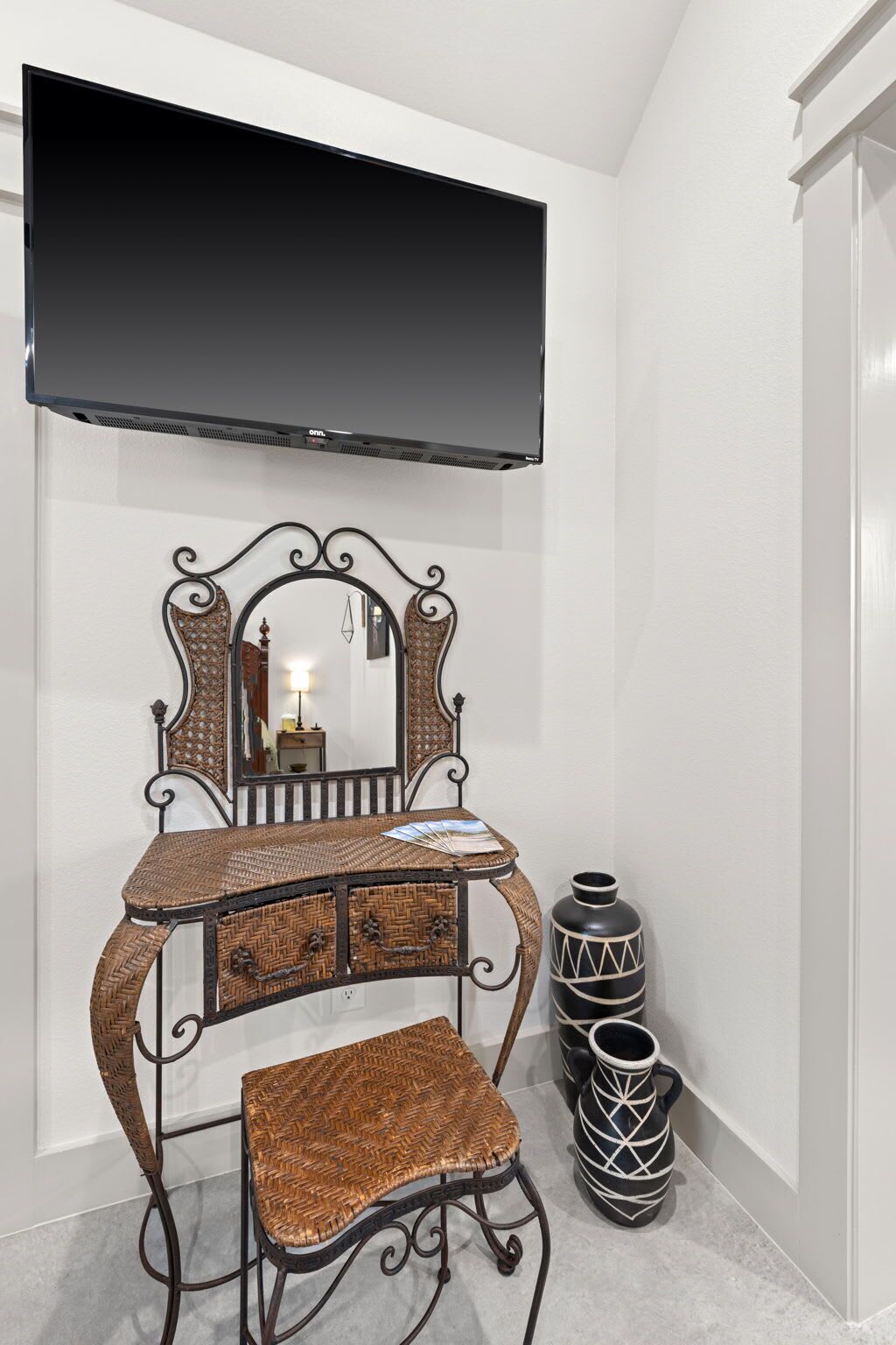A room with a television mounted on the wall, a decorative mirror, a wicker chair, and a black vase with geometric patterns.