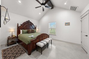 A bedroom with a wooden bed, a bench, a ceiling fan, a window with blinds, and a door. - Vista Moon: Cabin I - Pool, Chapel, Fire Pit (Fredericksburg)