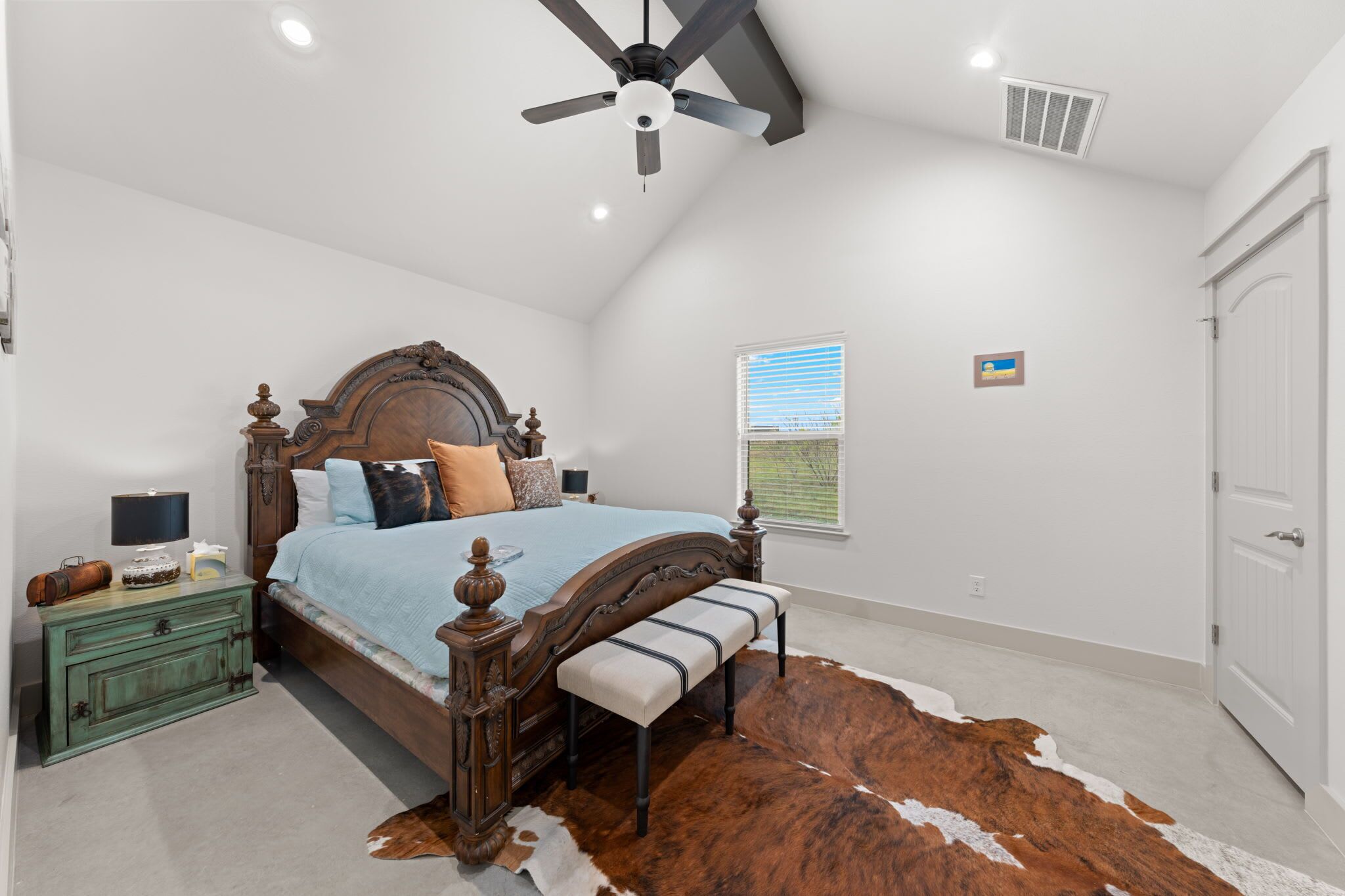 Une chambre à coucher avec un lit en bois, un ventilateur de plafond, une fenêtre avec des stores, une porte et un tapis en peau de vache.