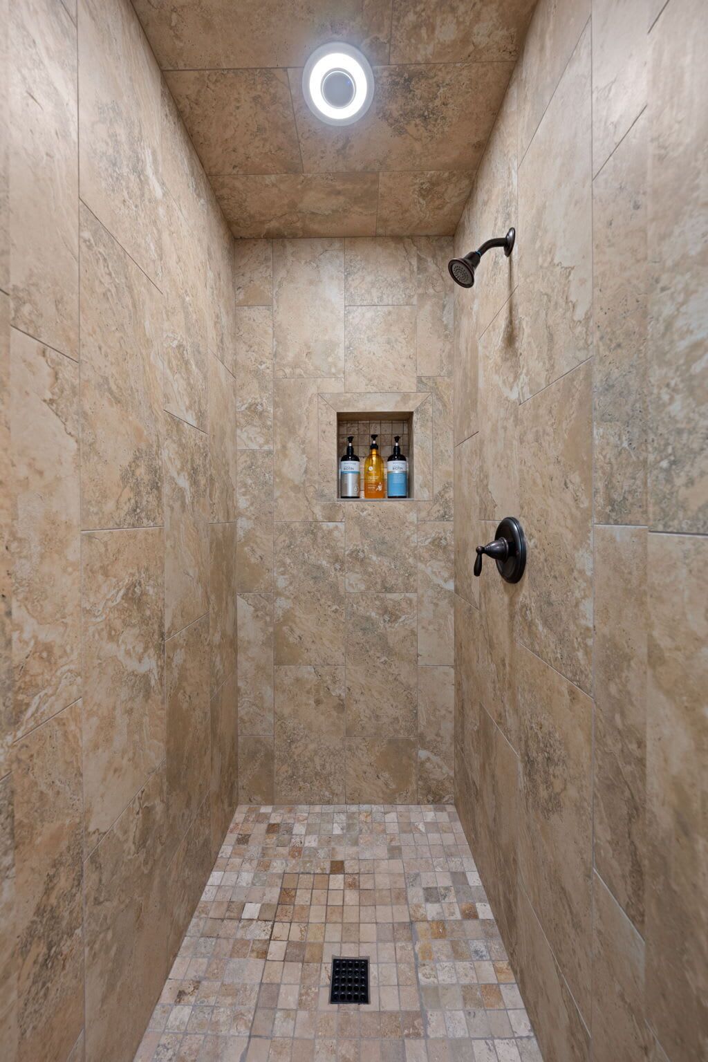 A narrow shower with beige tiles, a recessed ceiling light, and a small shelf with toiletries.