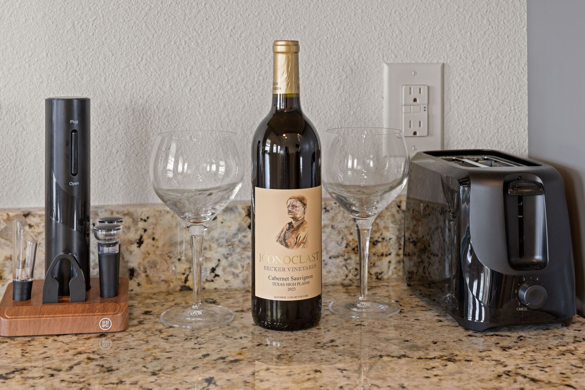 Una botella de vino, dos copas, un tostador y una cafetera sobre una encimera de cocina.