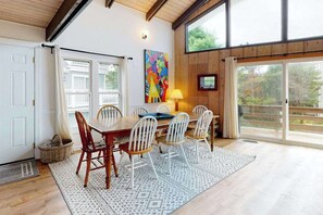 A dining area with a wooden table and chairs, a colorful abstract painting, and a view through large windows.