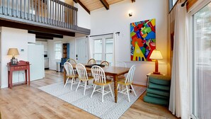 A dining area with a wooden table and white chairs, a colorful abstract painting on the wall, and a balcony with a railing.